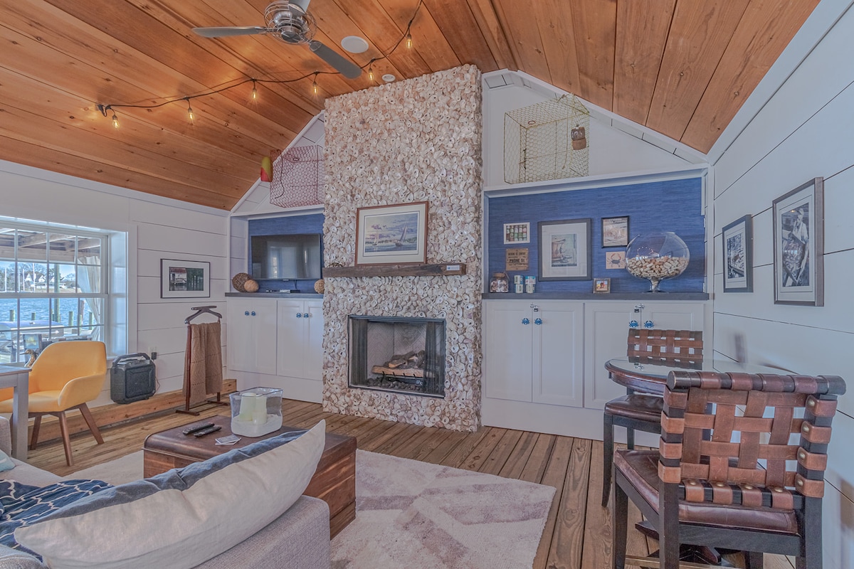 A cozy living area features a distinctive floor-to-ceiling oyster shell fireplace surrounded by wood paneling. Light-toned wooden beams create an inviting ambiance. Comfortable seating is arranged on a textured area rug, with framed artwork and nautical decor enhancing the space.