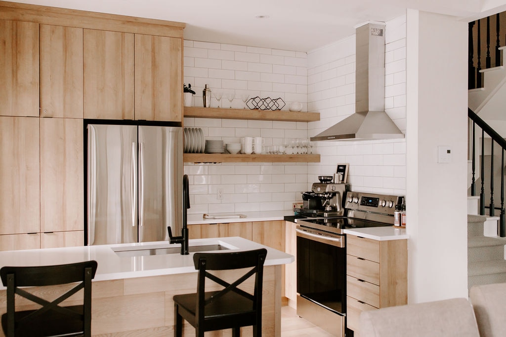 A modern kitchen features light wood cabinetry and white subway tile backsplash. Stainless steel appliances include a refrigerator and oven. An island with a sink offers additional counter space, complemented by two black chairs. Open shelving displays dishware above the countertop.