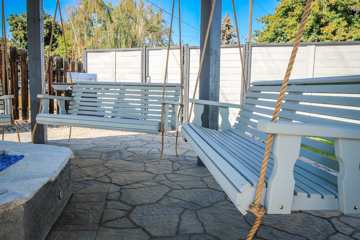 Two white wooden swings on a stone patio are positioned beneath a covered structure. A circular gas firepit is visible in the center, surrounded by a crafted stone surface. Lush greenery and a blue sky create a serene backdrop.