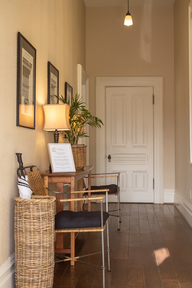 A well-lit hallway features warm wooden flooring and neutral walls. Framed artwork lines the walls, complemented by a lamp on a console table. A potted plant adds a touch of greenery, while a decorative basket and umbrella stand provide functional storage.