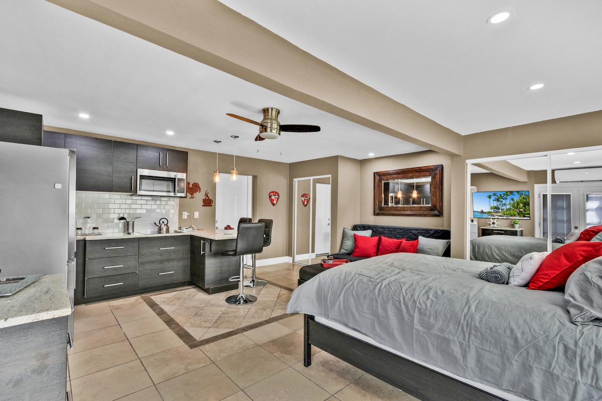 A modern studio layout is displayed, featuring a spacious bed with neutral bedding accented by red pillows. A sleek kitchen area with dark wood cabinets, stainless steel appliances, and a small dining area are visible, along with a comfortable seated area in the background.