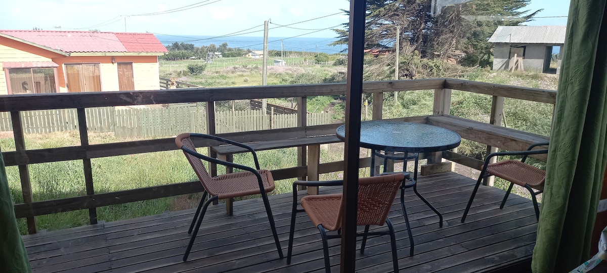 A wooden deck features a round table surrounded by two chairs, providing a casual outdoor seating area. Natural surroundings and distant views of the ocean are visible, creating a serene atmosphere. Light green curtains frame the view from inside, adding a touch of warmth.