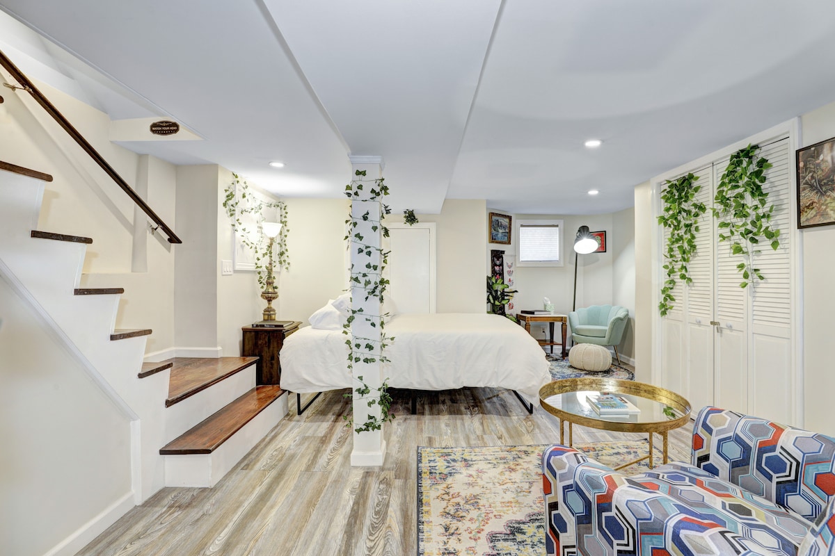 A garden level studio apartment features a king-sized bed with white linens centered in the room. A cozy reading chair sits next to a small side table, while colorful armchairs are positioned nearby. Recessed LED lighting brightens the space, complemented by decorative plants along the walls.