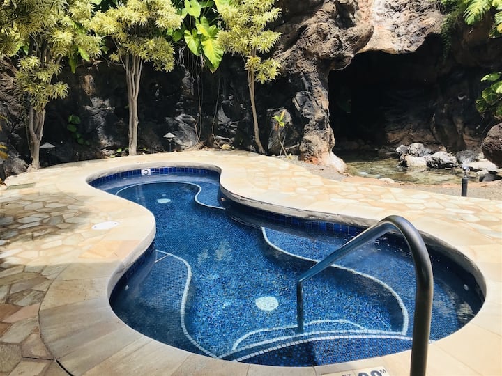 A curvy lagoon-style pool is surrounded by lush greenery, featuring smooth stones and a mosaic-tiled surface. The water reflects light, creating a serene atmosphere with a natural rock backdrop that enhances the tropical feel.