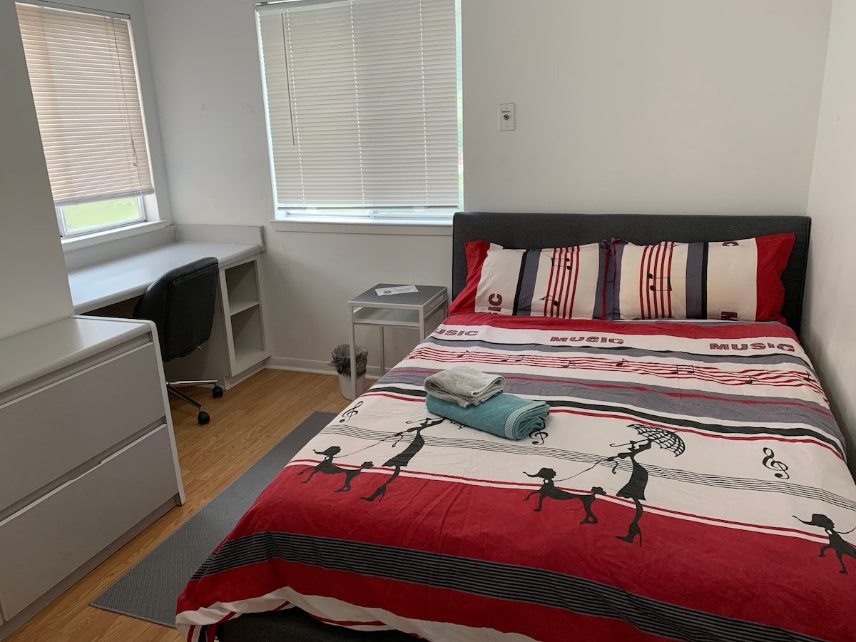 A well-lit room features a spacious bed covered with a striped red and white blanket. A study desk is positioned near a window, providing natural light. A dresser is visible, and towels are neatly placed on the bed, enhancing the room's functionality.
