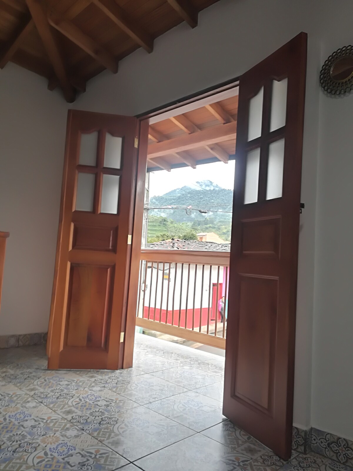 A pair of wooden doors stands open, revealing a scenic view of lush green mountains in the distance. The balcony railing adds an element of depth, while the interior highlights tiled flooring and the rustic wooden ceiling overhead.