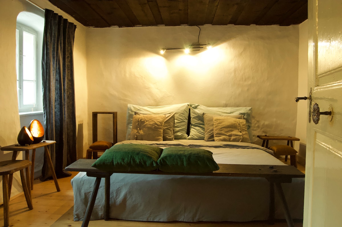 A sleeping area showcases a spacious bed with soft blue bedding and several cushions. A cozy bench is positioned at the foot of the bed, while two wooden stools provide additional seating. Warm lighting highlights the textured walls and wooden ceiling.