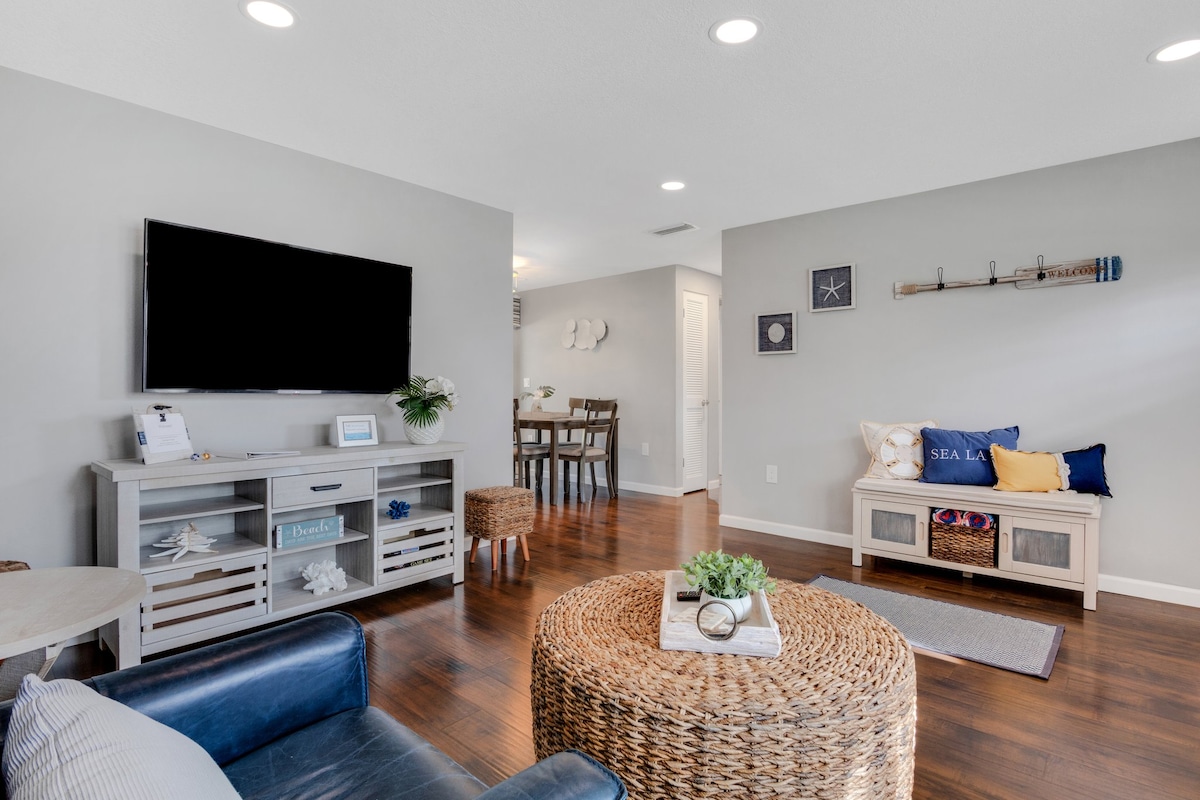 A living area is depicted with a flat-screen TV mounted on a neutral wall. A comfortable navy blue armchair faces a round woven coffee table. A small decorative piece rests atop the table, while a bright seating area features a light-colored bench adorned with blue and white cushions. Natural lighting enhances the space.