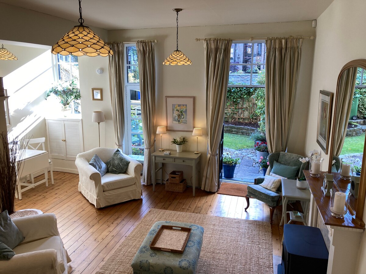 The living area features natural light streaming through large windows, illuminating soft seating and warm wooden floors. Two elegant light fixtures hang from the ceiling, and a small table sits between two armchairs. A view of the garden invites the outdoors inside.
