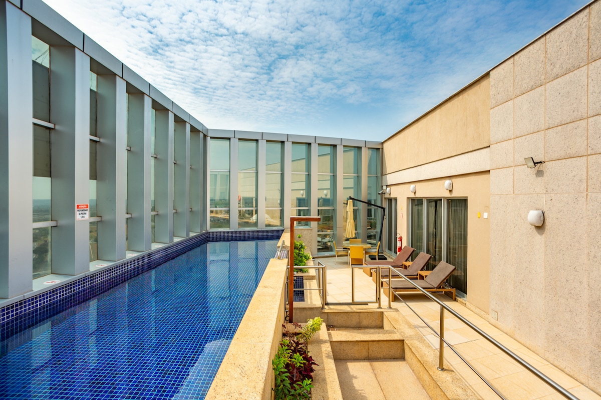 A tranquil rooftop area features a serene swimming pool with inviting blue water, surrounded by contemporary architecture. Sun loungers are placed nearby on a patio, providing a comfortable space for relaxation. The sky above is bright and partly cloudy, adding to the serene atmosphere.