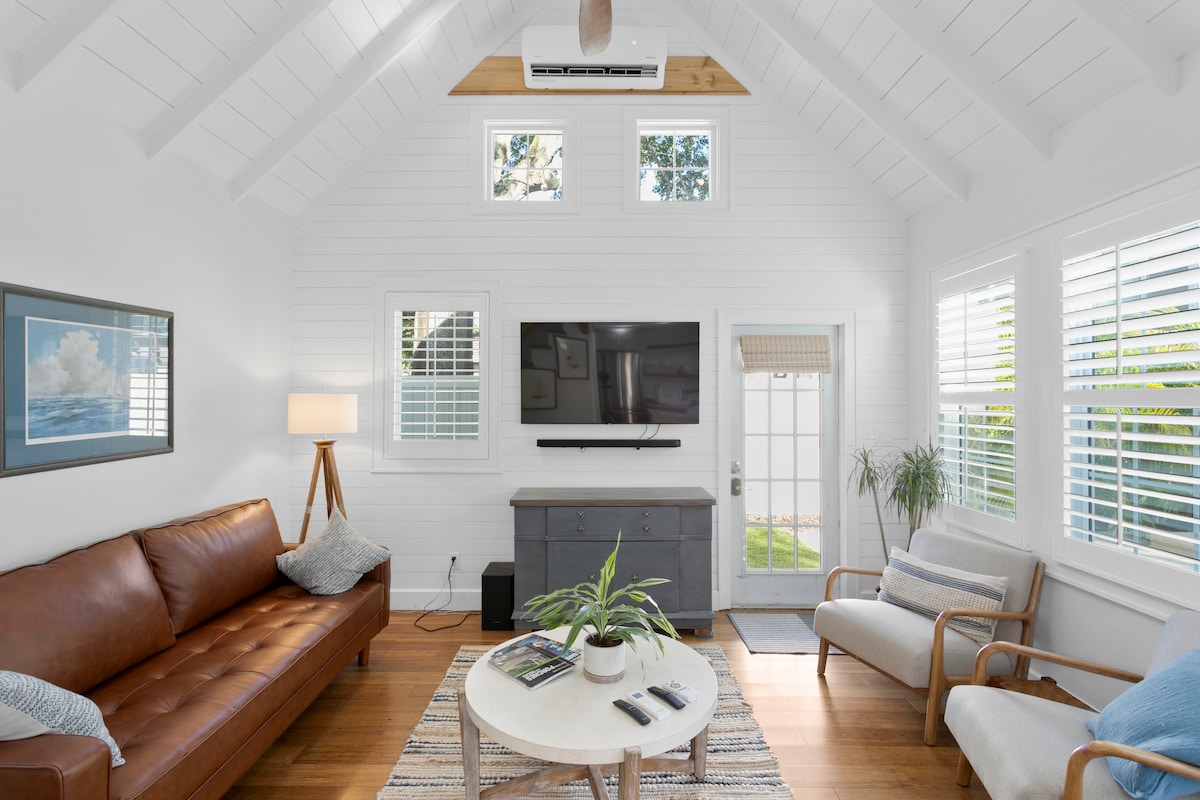 A bright living space features a brown leather sofa and light-colored accent chairs. A medium-sized coffee table is arranged with magazines. Natural light filters through window treatments, highlighting the white walls and wood flooring. A mounted television is centered on the wall, enhancing the open atmosphere.