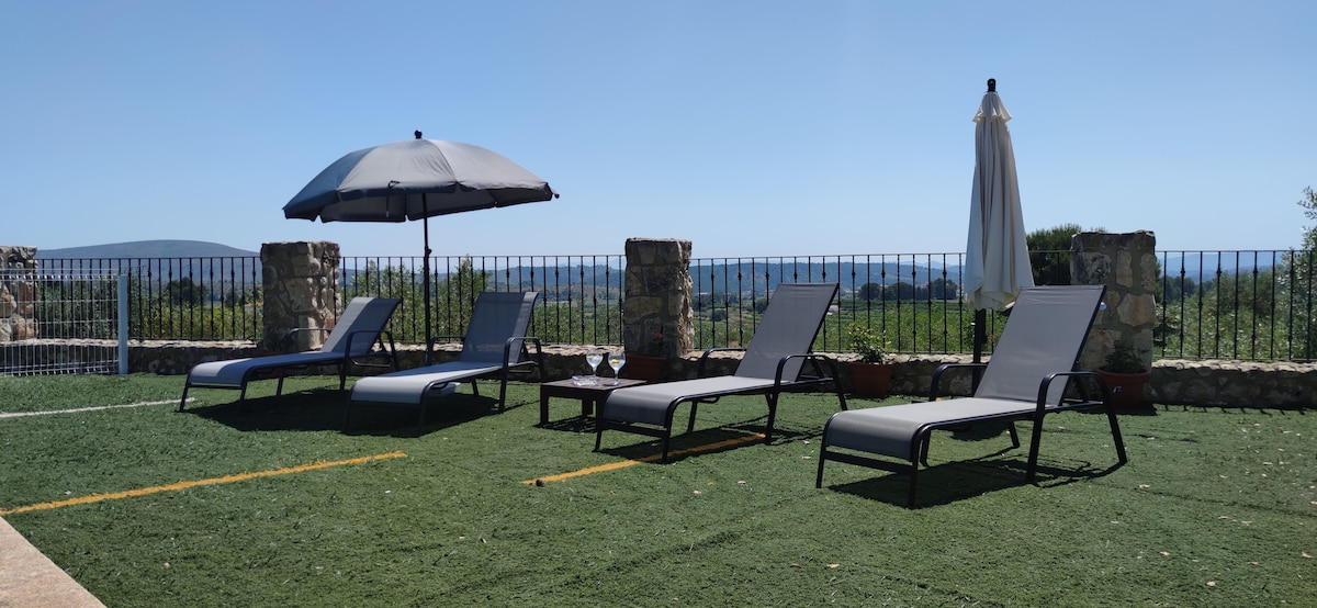 Lounge chairs are arranged on a green lawn area, accompanied by a large umbrella for shade. A small table is positioned nearby, creating a space for relaxation. In the background, open views of the surrounding landscape and sky are visible, providing a serene atmosphere.