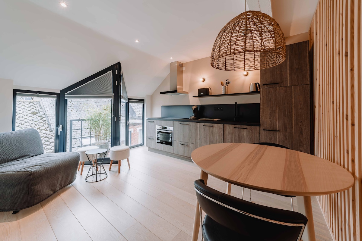 A modern living area features a comfortable gray sofa and two small tables, accompanied by a dining table with black chairs. A well-equipped kitchen with wood and dark cabinetry is visible, complemented by large windows allowing natural light to brighten the space.