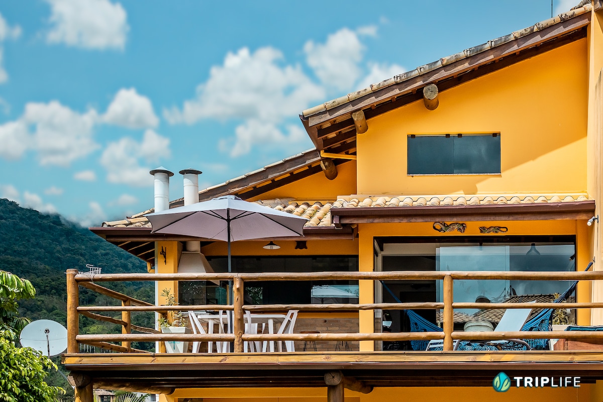 The exterior of the vibrant yellow house features a wooden deck with an outdoor umbrella and several chairs. Large windows provide an unobstructed view of the surrounding landscape. The roof is adorned with terracotta tiles, and the mountains are visible in the background.