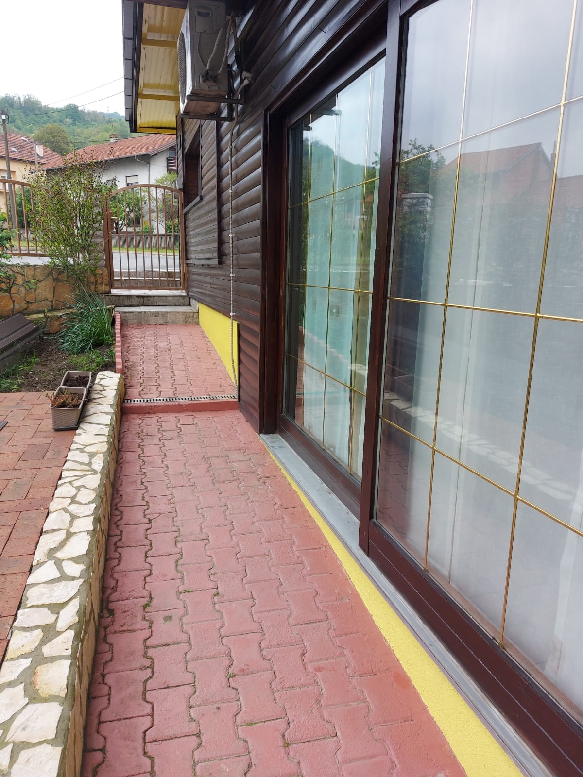 A paved walkway leads to the entrance of the accommodation, bordered by a low stone wall and greenery. Large glass doors offer a view into the interior, framed by a wooden exterior. The pathway is lined with rectangular bricks, enhancing the outdoor area.