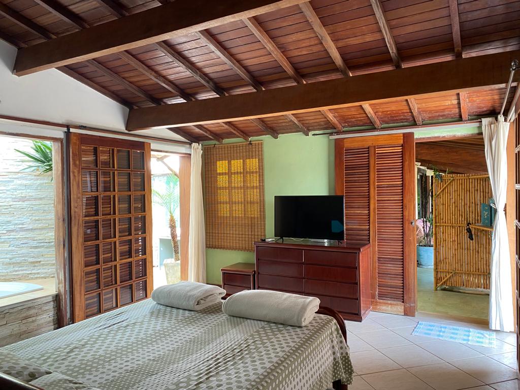 A spacious sleeping area features a wooden ceiling and is accompanied by a comfortable bed. A television rests on a dark wooden dresser, while large windows allow natural light to illuminate the room. A bamboo screen adds a touch of privacy and warmth.