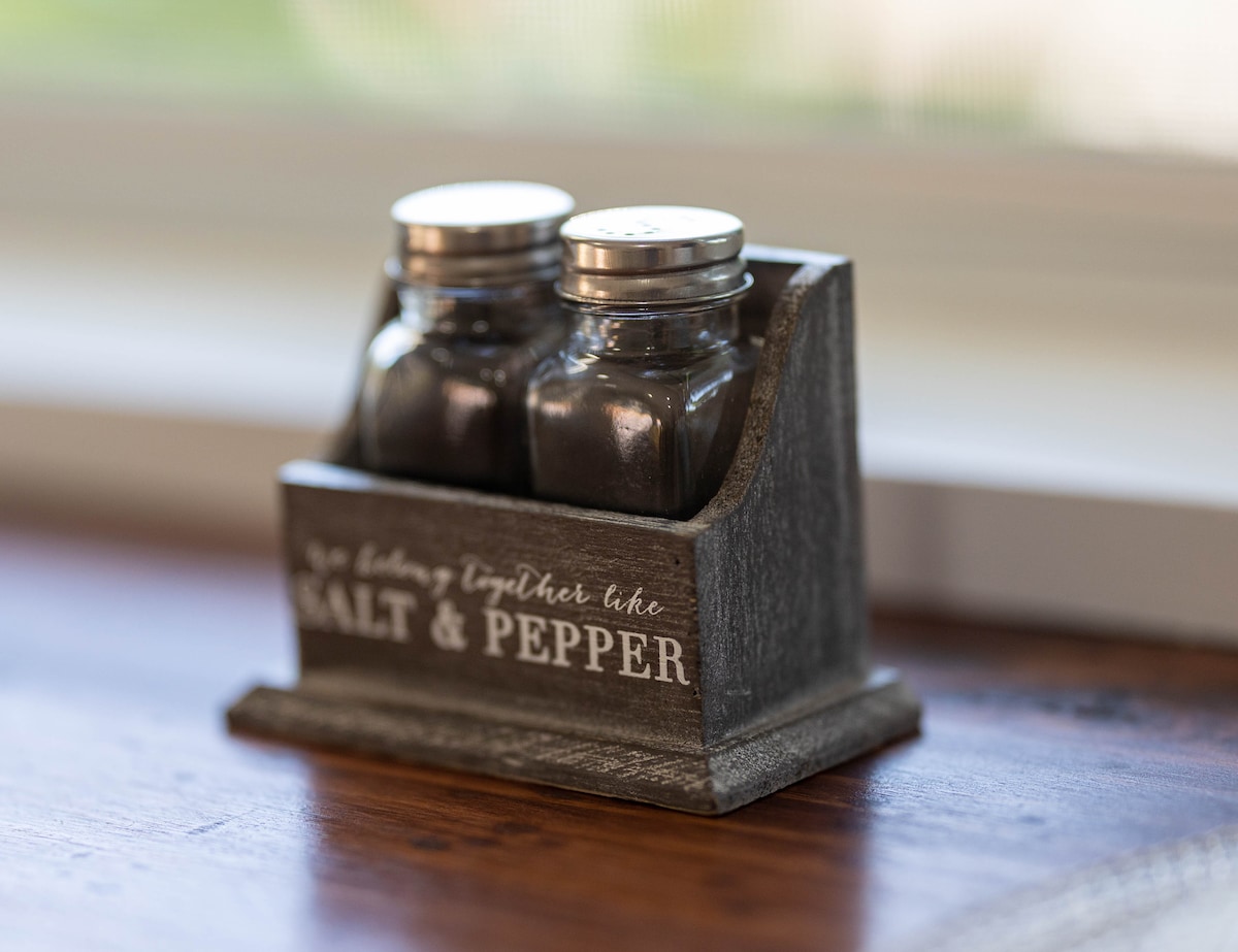 A rustic salt and pepper shaker set is displayed on a wooden surface. The shakers are glass with metal lids, housed in a stylish wooden holder featuring a decorative text design. Natural light softly illuminates the set, creating a warm, inviting atmosphere.