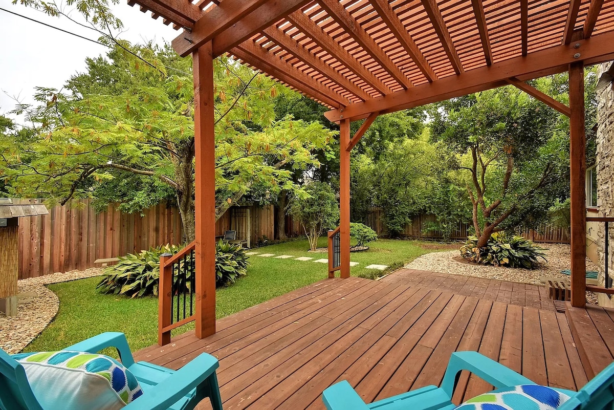 A wooden deck is framed by a pergola, offering seating on bright turquoise chairs. Lush greenery surrounds the outdoor space, featuring trees, shrubs, and a neatly maintained lawn. Pathways of soft stone lead through the garden area, creating a serene retreat.