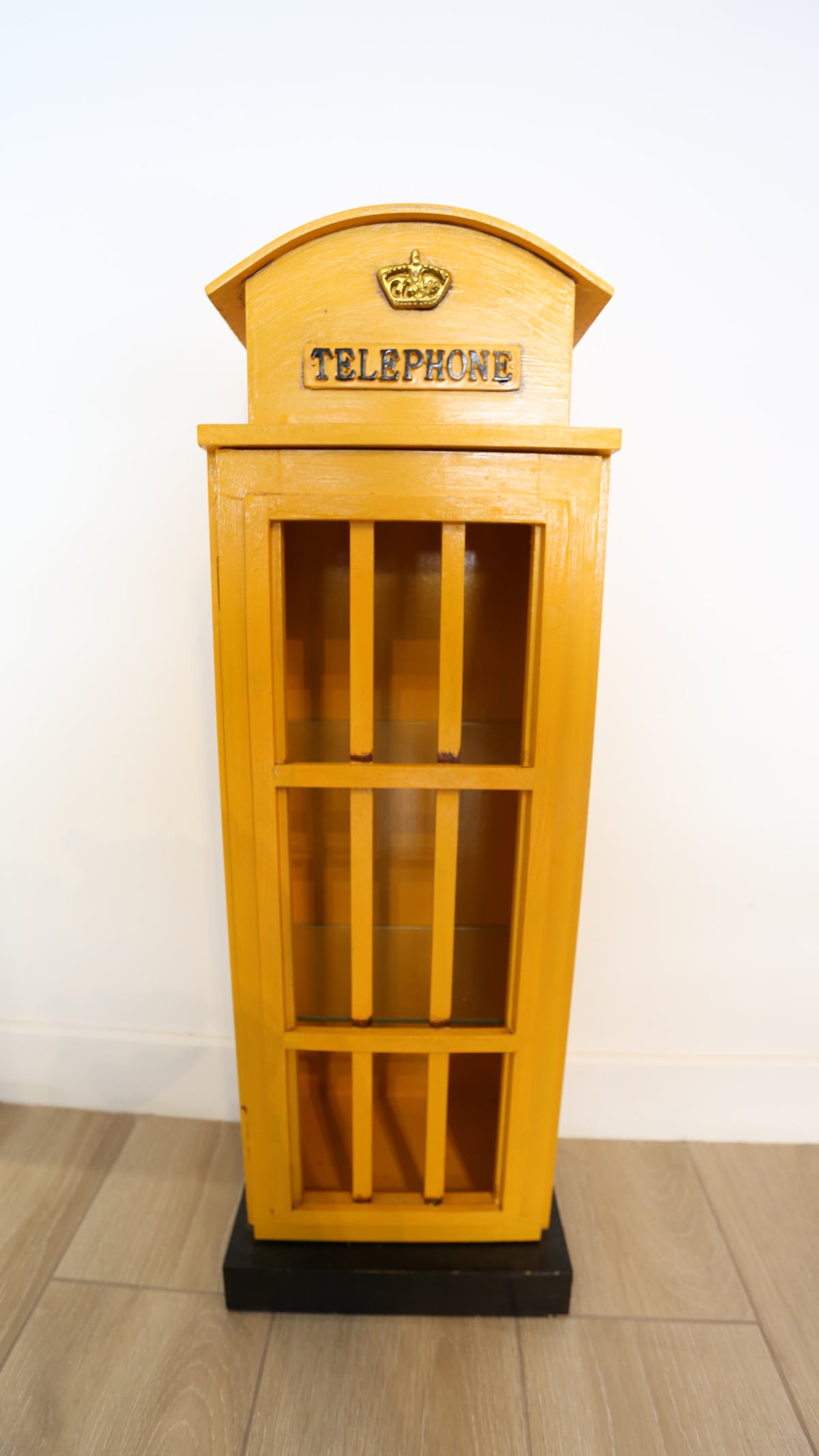 A vintage-style telephone booth stands upright, painted in bright yellow. The booth features a sign that reads 'TELEPHONE' and displays four rectangular windows on each side, allowing for an open view of the interior space.