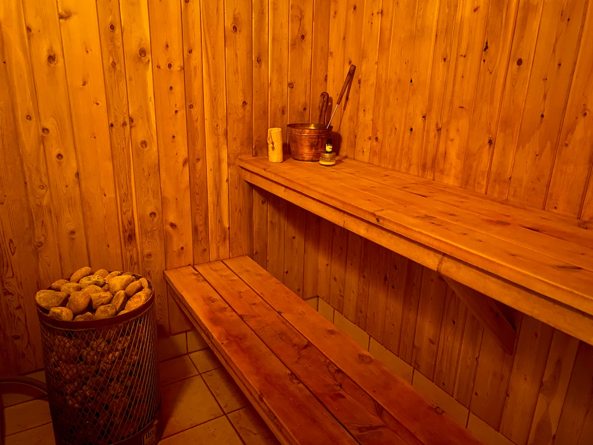 A warm wooden sauna interior features natural wood paneling and a spacious bench along the wall. A bucket and ladle are placed on a shelf, while a basket filled with smooth stones sits nearby, creating a serene and inviting space for relaxation.