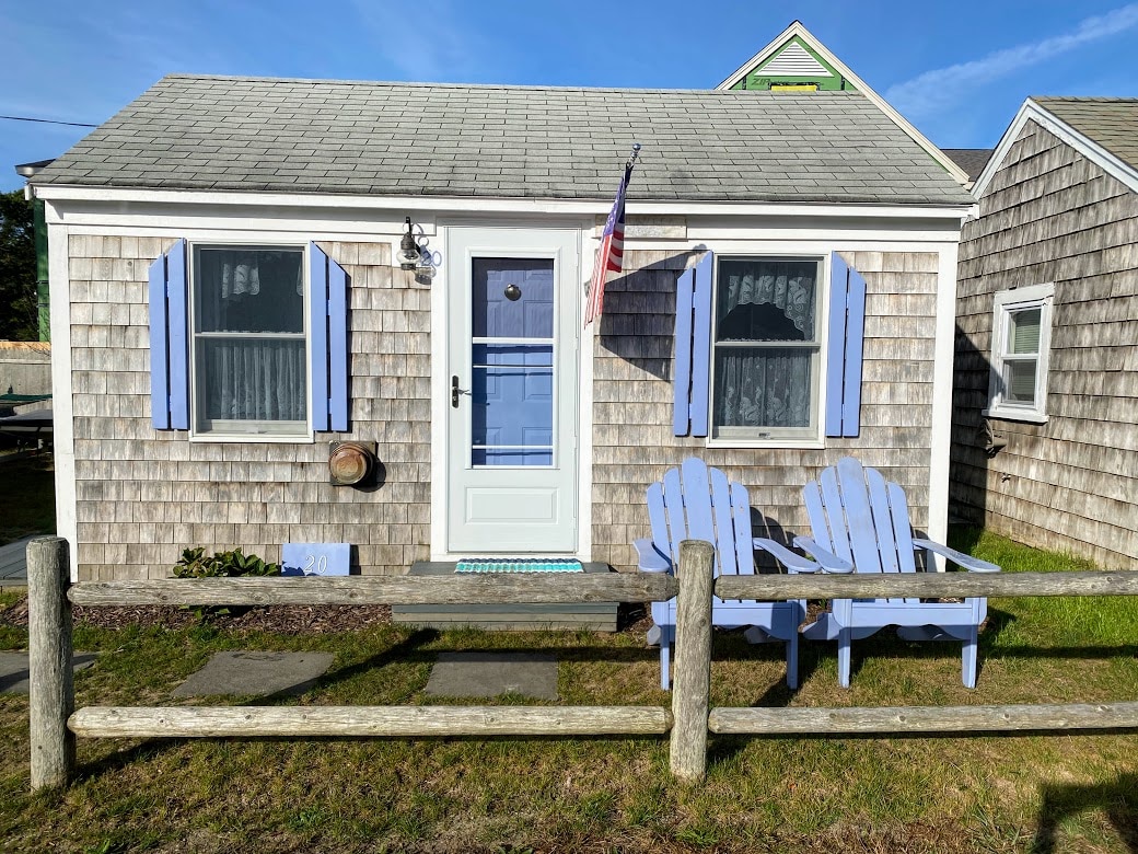 The exterior of a cozy cottage features a light blue door and matching shutters. Two adirondack chairs sit on a grassy area in front of the home, with a wooden fence partially visible. A flag can be seen hanging beside the entrance.