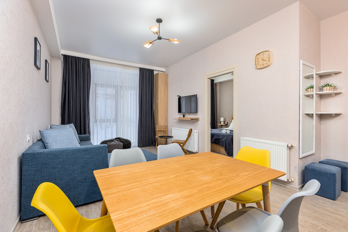 A contemporary living space is featured, showcasing a wooden dining table surrounded by colorful chairs. A spacious blue couch sits nearby, complemented by a large window with sheer curtains, allowing natural light to fill the room. A wall-mounted TV is visible, along with a decorative mirror and shelves.