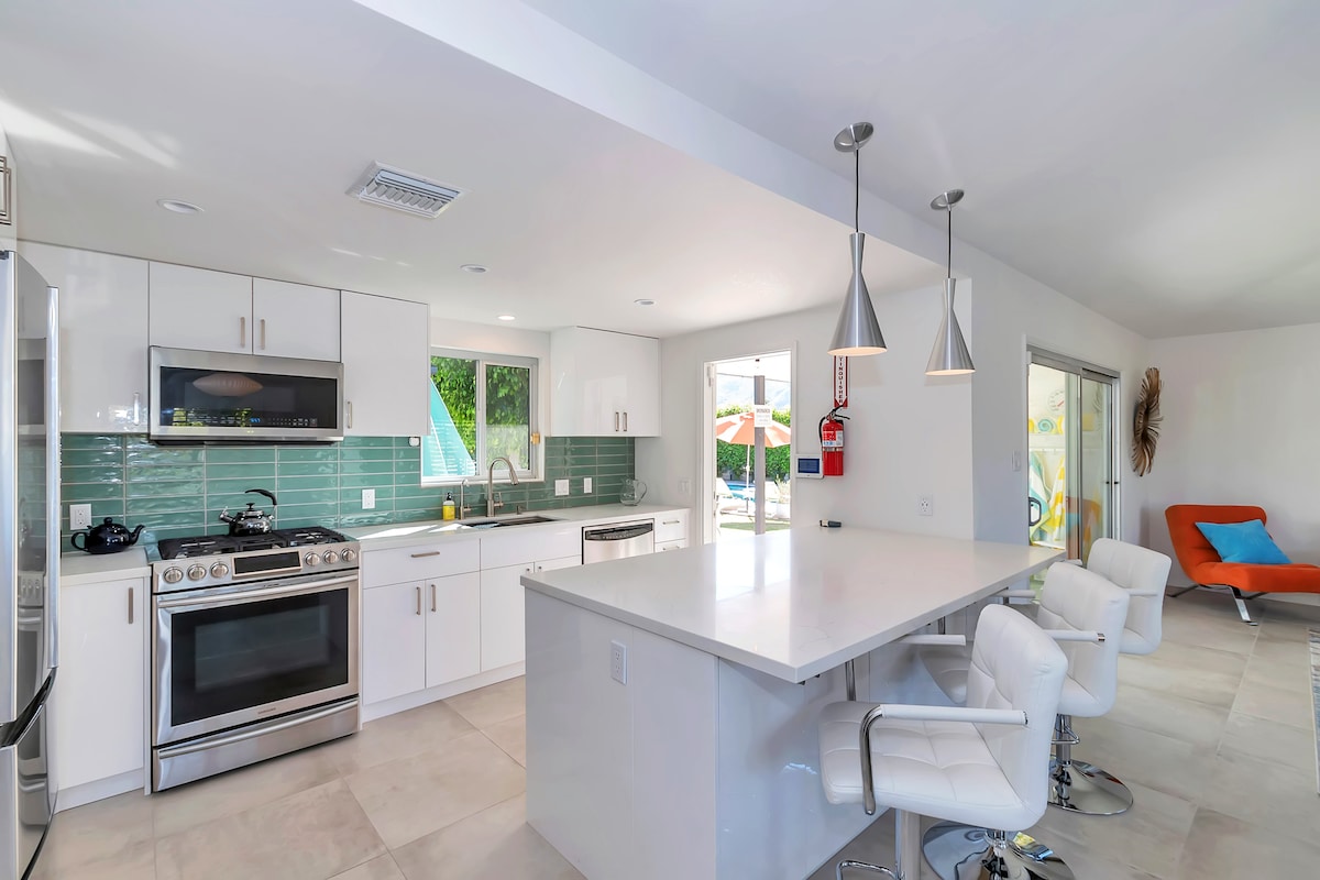 A modern kitchen is equipped with stainless steel appliances and a spacious island featuring bar stool seating. Natural light floods the area through large windows, illuminating the sleek white cabinetry and turquoise backsplash. The adjoining living space is visible in the background, enhancing the open layout.