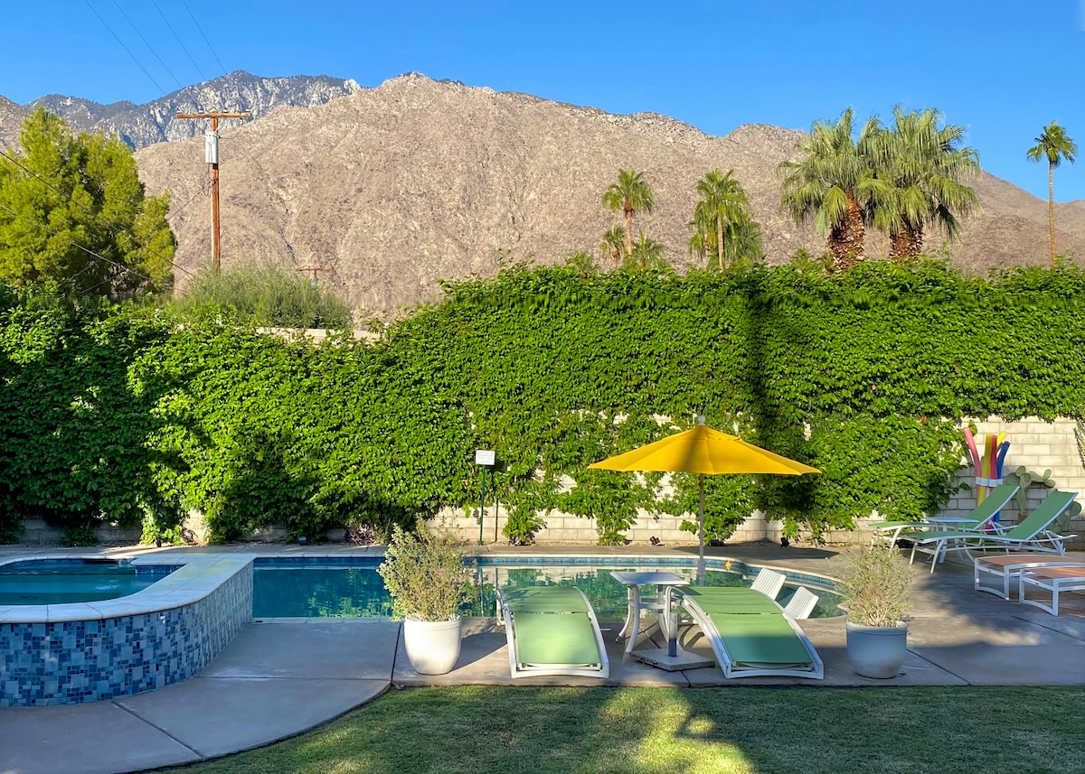 A large salt water pool is flanked by two lounge chairs, offering relaxation under the sun. A vibrant yellow umbrella provides shade. Lush greenery surrounds the area, complemented by a backdrop of impressive mountains and palm trees, creating a serene outdoor atmosphere.