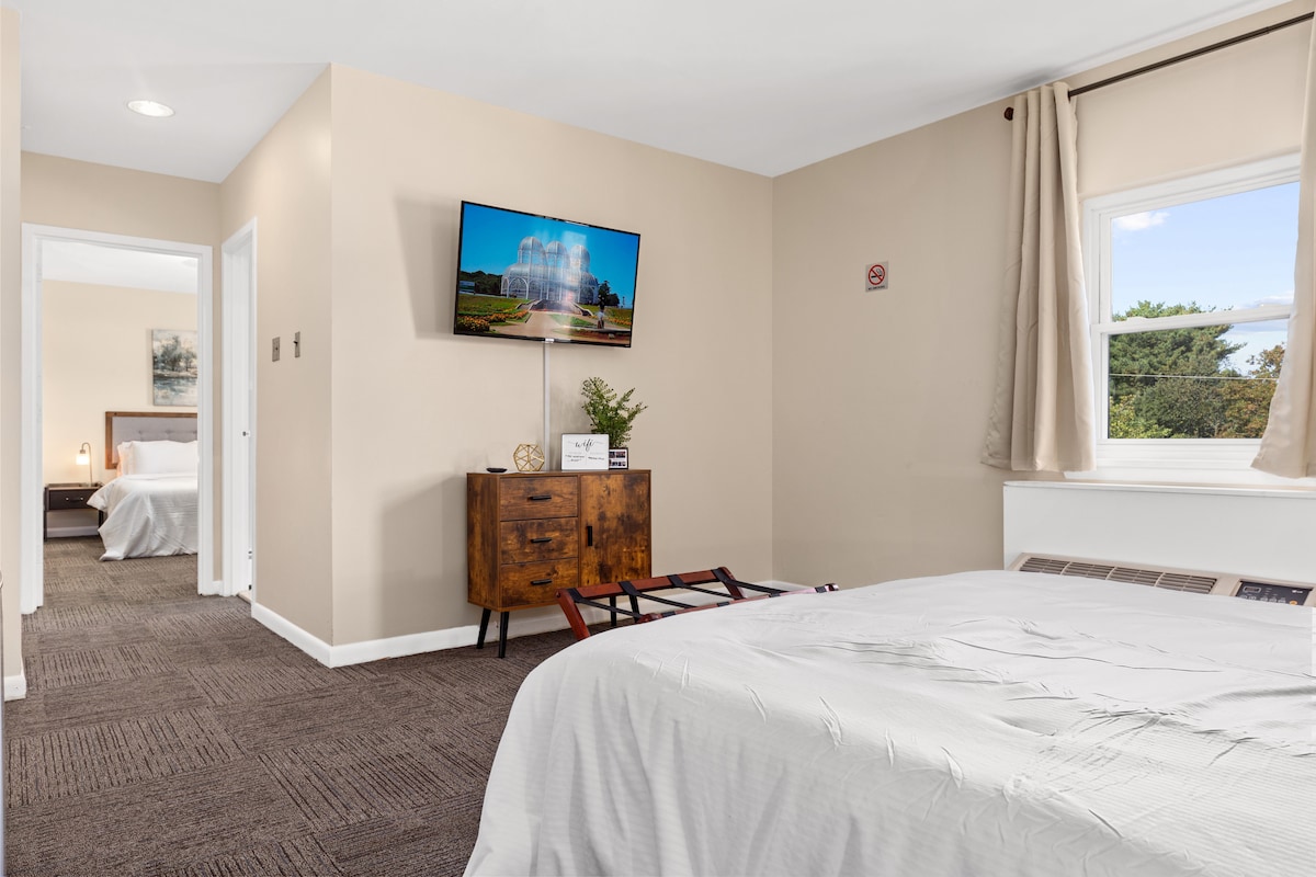A well-lit room is depicted with a spacious bed featuring crisp white bedding. A wooden dresser stands against the wall, supporting a flat-screen TV. Two doors are visible, leading to additional rooms, while a large window allows natural light to brighten the space.