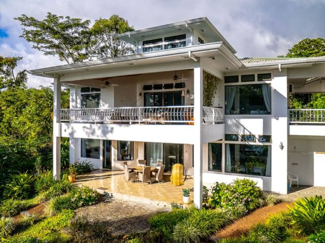 The exterior of the villa showcases a two-story structure surrounded by lush greenery. A spacious patio is visible, furnished with a dining set and decorative elements. Large glass doors provide an inviting entrance, while the upper balcony offers additional seating, enhancing the connection with nature.
