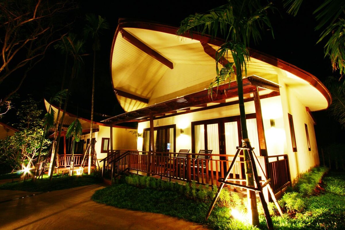 The bungalow's exterior is illuminated at night, showcasing a modern design with a curved roof. Wooden railings frame the porch, where several chairs are arranged. Lush greenery surrounds the building, adding to the serene atmosphere.