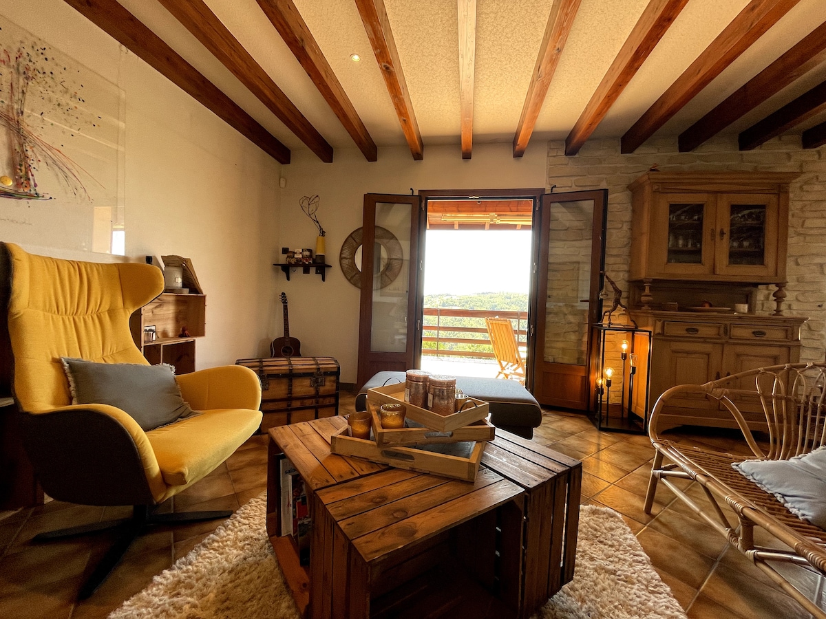 A cozy living area features a yellow armchair facing a wooden coffee table adorned with books and decor. Exposed wooden beams run across the ceiling, complementing the stone wall. Double doors open to a panoramic view, enhancing the inviting atmosphere of the space.