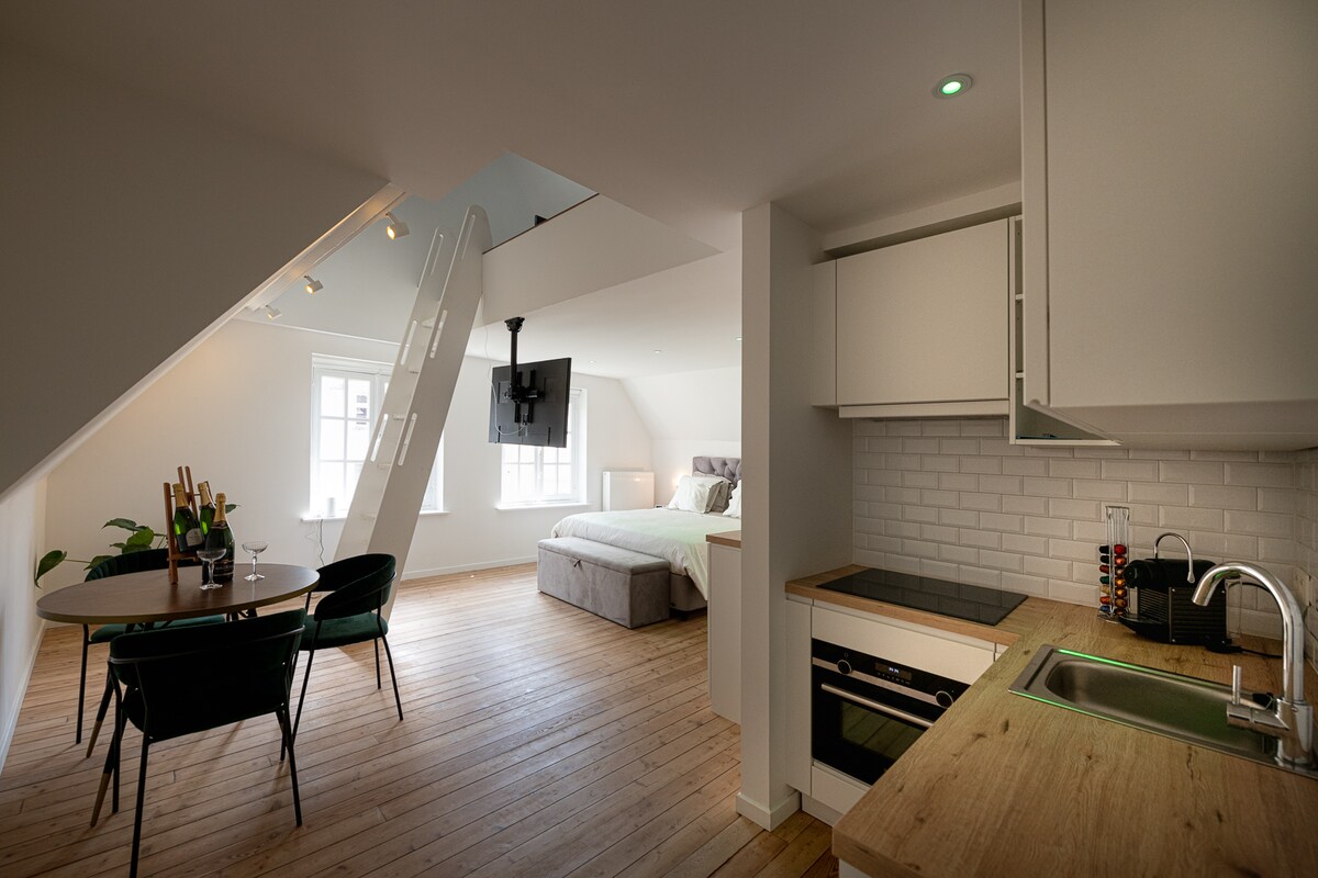 A modern suite features an open layout with a kitchenette and dining area. A round table with green chairs is positioned next to a spacious bed, while natural light floods the room through several windows. Light wooden flooring complements the neutral decor.