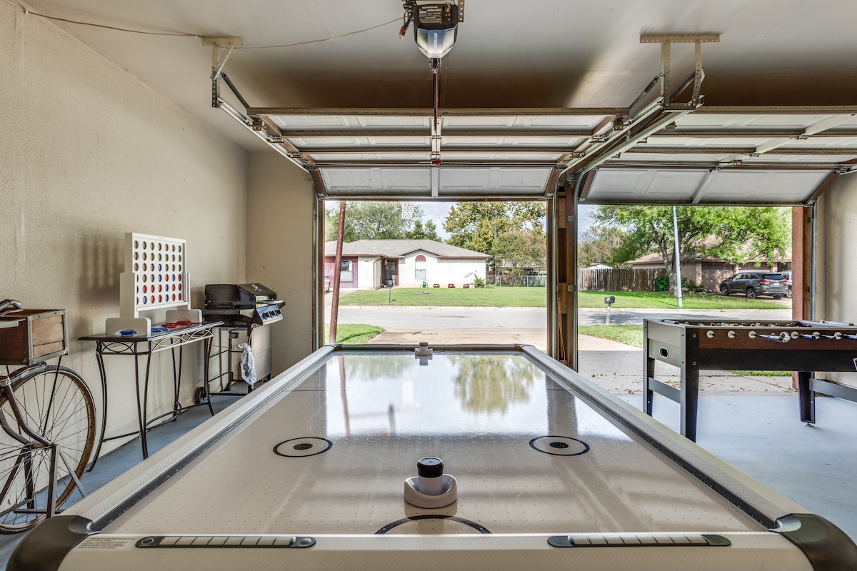 The garage features a large air hockey table as the main attraction, with a smooth surface ready for play. A barbecue grill and a small table with chairs are visible, providing additional space for leisure activities. Natural light enters from the open garage doors, illuminating the area.