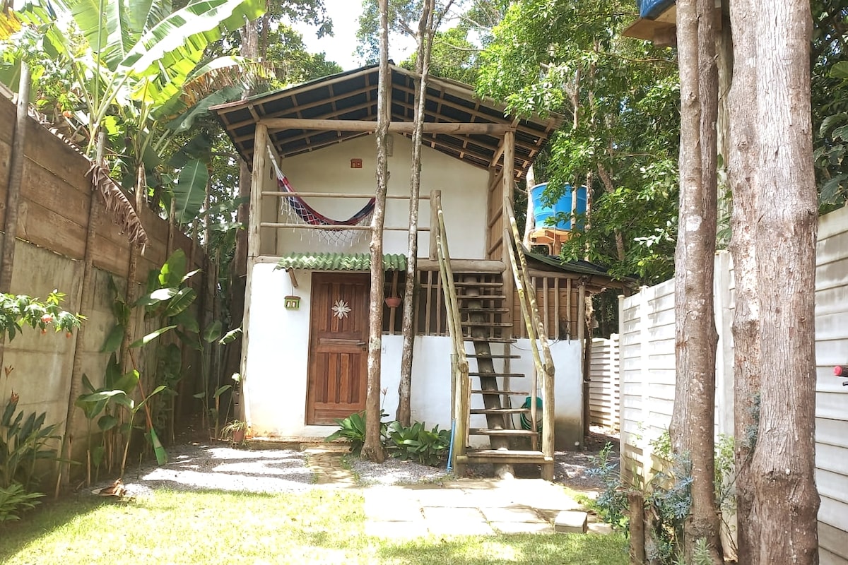A charming two-story bungalow is nestled among lush greenery. The structure features a wooden door and a staircase leading to a balcony. A hammock is visible on the upper level, surrounded by tropical plants, enhancing the serene environment.