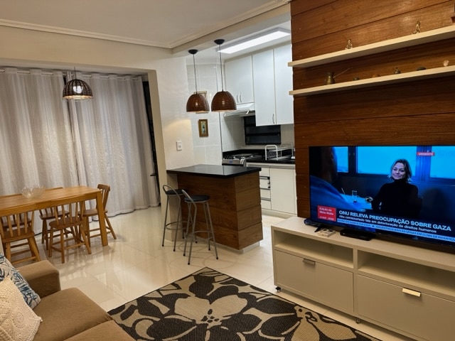 The open-plan living area displays a dining table with six chairs beside a modern kitchen. A bar counter with stools separates the kitchen and living space. A large TV is positioned on a sleek entertainment unit, contributing to a functional and inviting environment.