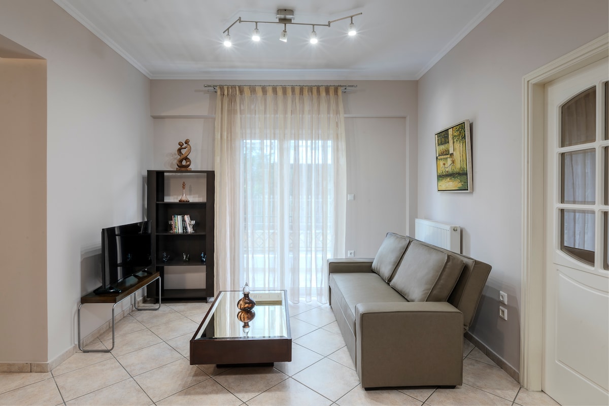 A comfortable living room features a modern sofa and a low coffee table. A television is positioned on a sleek stand. Natural light fills the space through large glass doors draped with sheer curtains. A decorative piece rests on the table, enhancing the room's warmth.