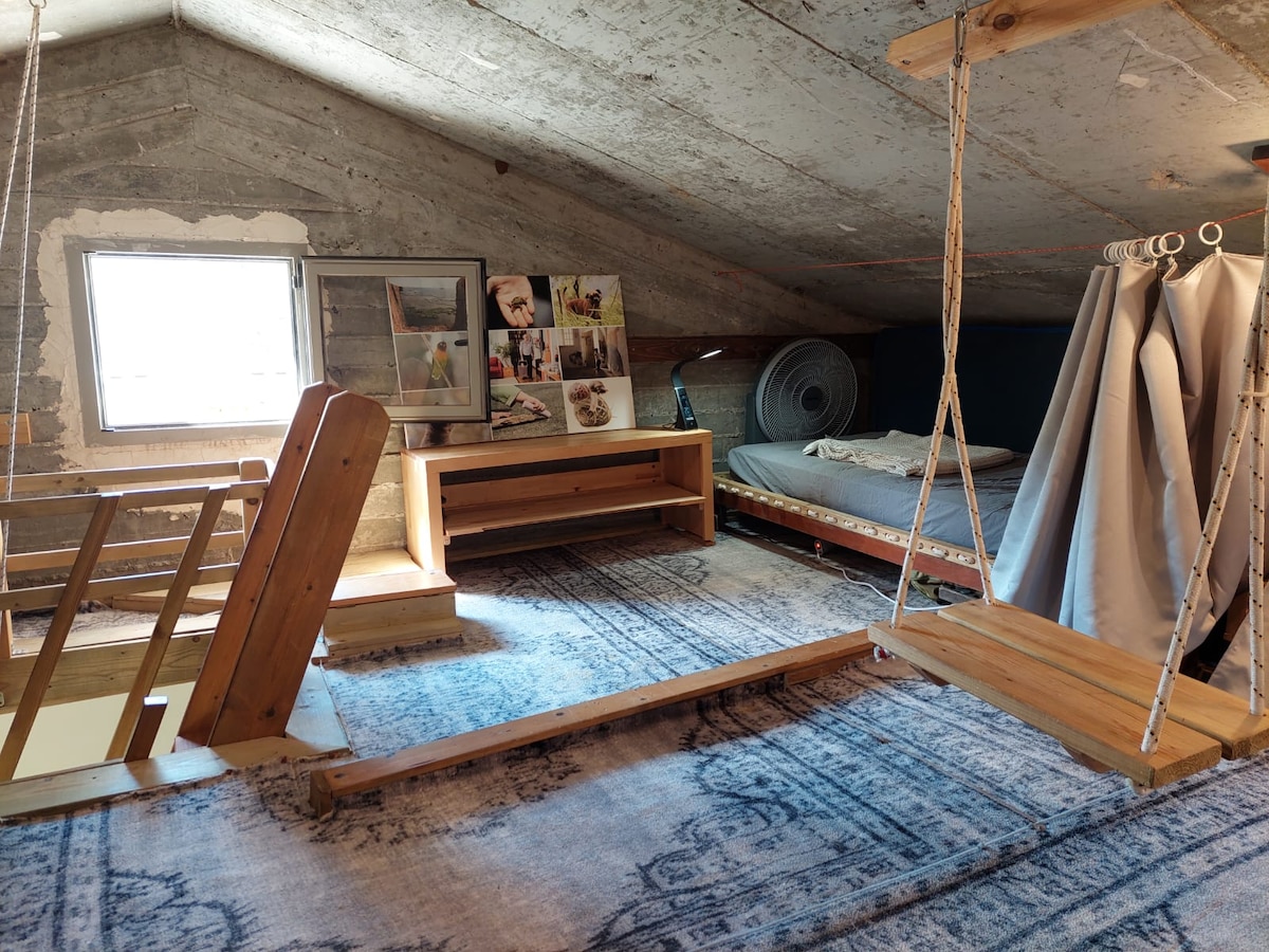 The spacious attic is adorned with wood accents and features a comfortable sleeping area with two mattresses. A window allows natural light to enter, while a rustic wooden swing adds unique charm. Decorative artwork is displayed on the wall, enhancing the inviting atmosphere.