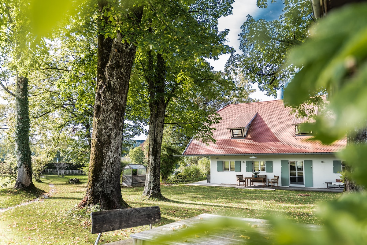 A charming house is nestled among tall trees, featuring a red sloped roof and large windows. A spacious patio with outdoor seating is visible, surrounded by a well-maintained garden. Pathways lead through the serene landscape, inviting exploration.