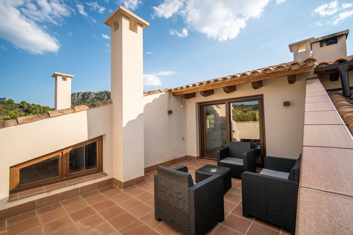 A cozy terrace area is visible, featuring two outdoor chairs and a small table positioned on tiled flooring. The backdrop showcases a clear sky, and the stone chimney complements the modern design of the structure.