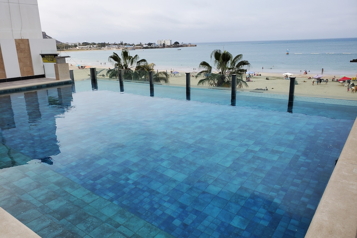 An infinity pool is visible, overlooking the beach with ocean waves gently lapping at the shore. Towering palm trees frame the edges of the pool, and the water reflects various shades of blue against a backdrop of a cloudy sky.