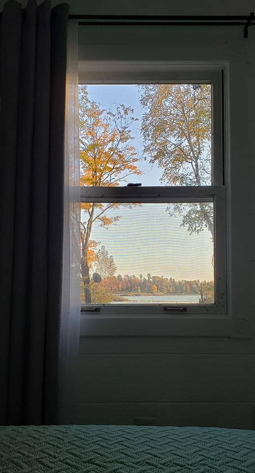 A window frame showcases a view of trees displaying autumn colors against a tranquil lake backdrop. Soft natural light filters through sheer curtains, highlighting the peaceful atmosphere within the Tiny House.