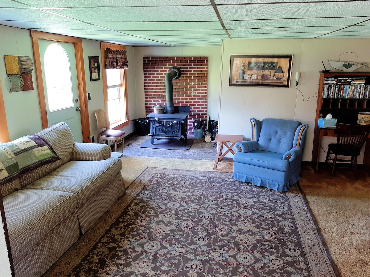 A cozy living area features a comfortable couch and an armchair, both positioned around a brick fireplace. A wooden table and chairs are present, along with a bookcase displaying various items. Natural light enters through multiple windows, creating a warm atmosphere.