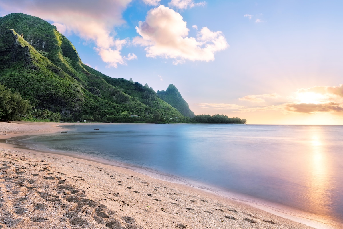 A serene beach scene showcases soft sand with gentle footprints leading towards calm ocean waters. Lush green mountains rise in the background, reflecting the soft light of the setting sun, creating a tranquil coastal atmosphere.