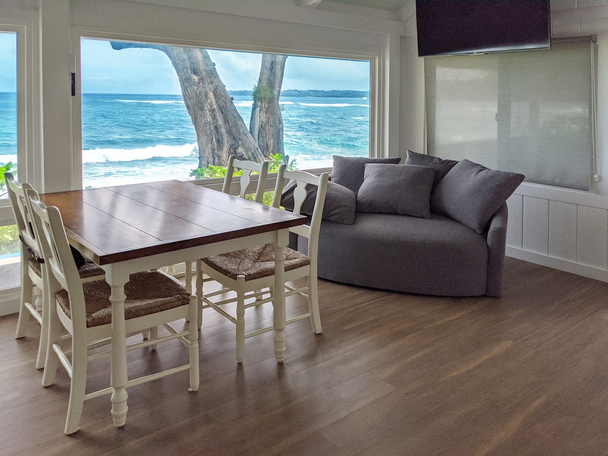 A spacious dining area features a large wooden table surrounded by white chairs. Natural light floods the room through expansive windows overlooking the ocean. A cozy grey couch is positioned nearby, inviting relaxation while enjoying the scenic view of the coastline.