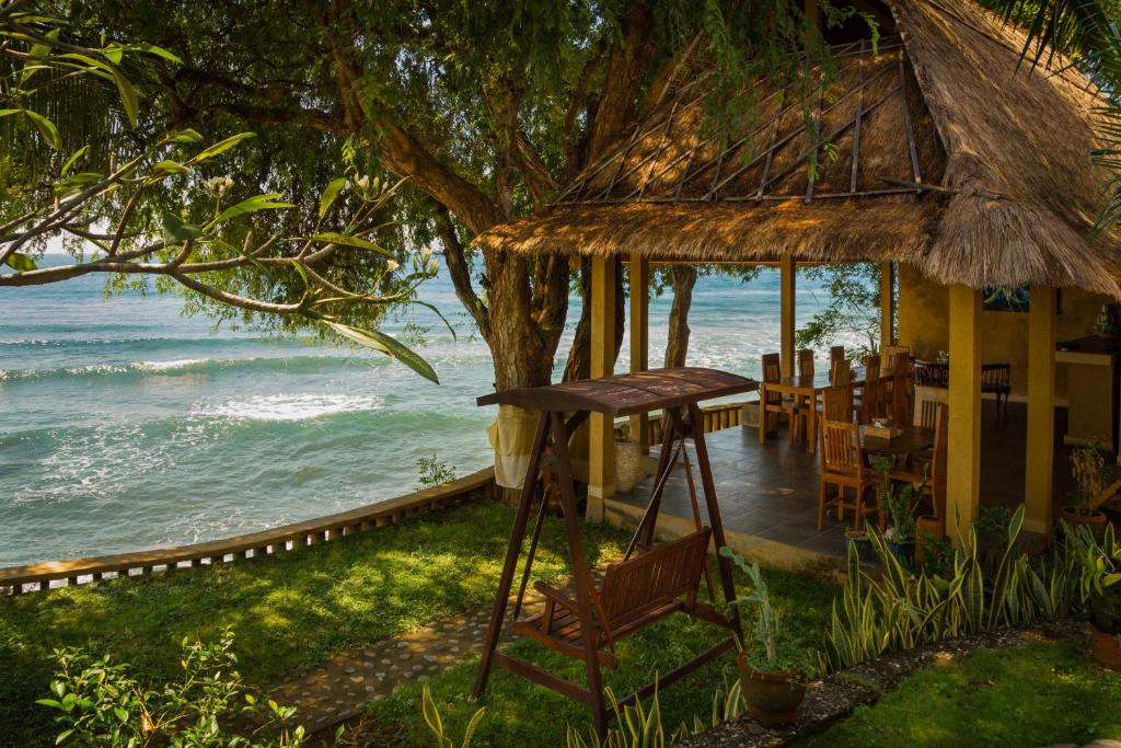 A swing set is positioned beside a grassy area, offering views of gently crashing ocean waves. A thatched-roof structure with wooden furniture is visible in the background, framed by lush greenery and trees providing shade.