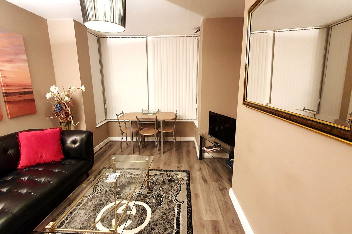 A modern living room features a black leather sofa with a pink cushion, complemented by a glass coffee table. A dining table with four chairs is set against a large window, while a wall-mounted TV is positioned nearby. Neutral-toned walls create a spacious ambiance.