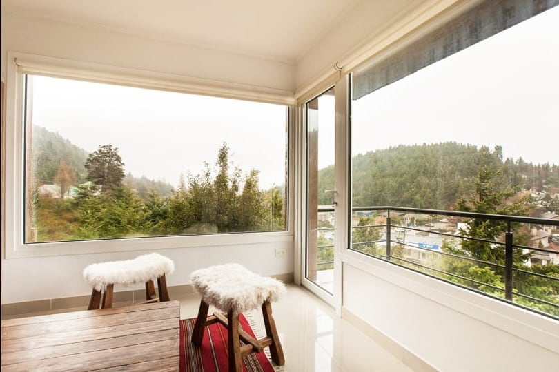 A bright corner of the room features large windows that offer expansive views of the surrounding greenery. Two wooden stools with cozy white coverings are positioned around a low wooden table, enhancing the inviting atmosphere of the airy space.