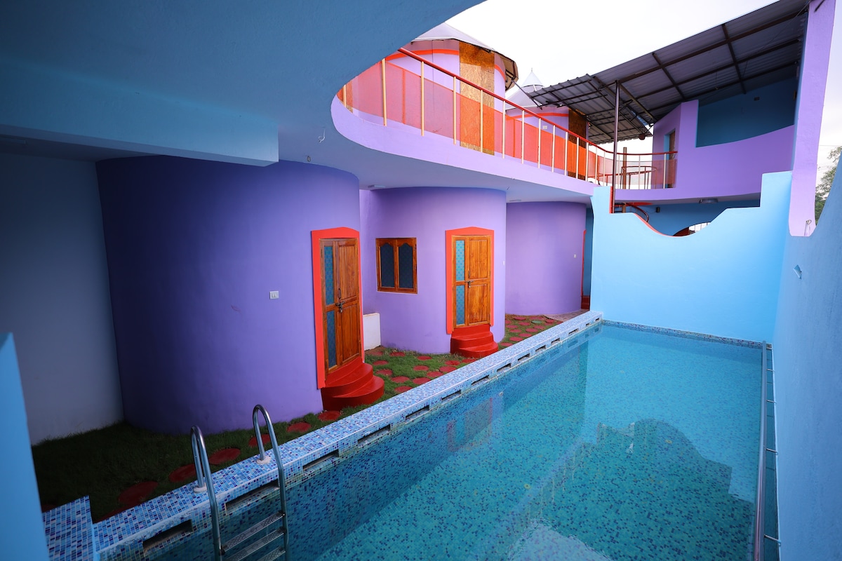 An outdoor area features a vibrant blue indoor swimming pool surrounded by smooth tiles. Colorful round rooms with orange doors are situated nearby, complemented by a green lawn. A covered area above offers shade, providing a tranquil space for relaxation.