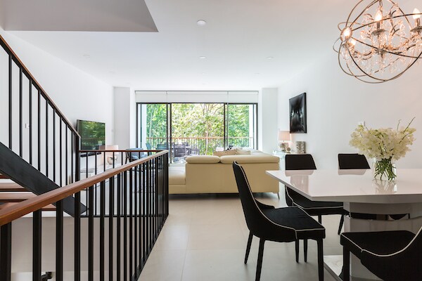 An open living area is highlighted by a sleek design, featuring a large sectional sofa and a flat-screen TV. A contemporary dining table with dark chairs is positioned near a glass door, which provides views of the lush greenery outside.